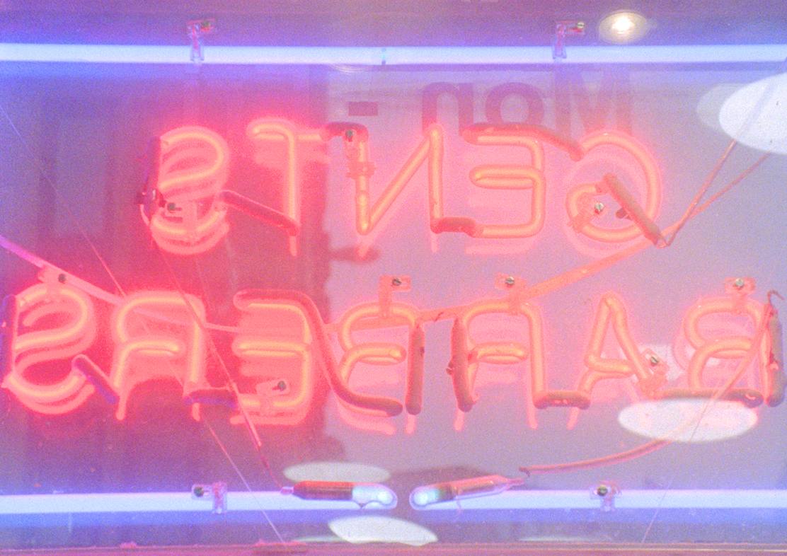 An image of a red neon light sign on a blue square outline, which reads -gents barbers- inverted, as it was photographed from the inside of the shop (the sign points outwards)